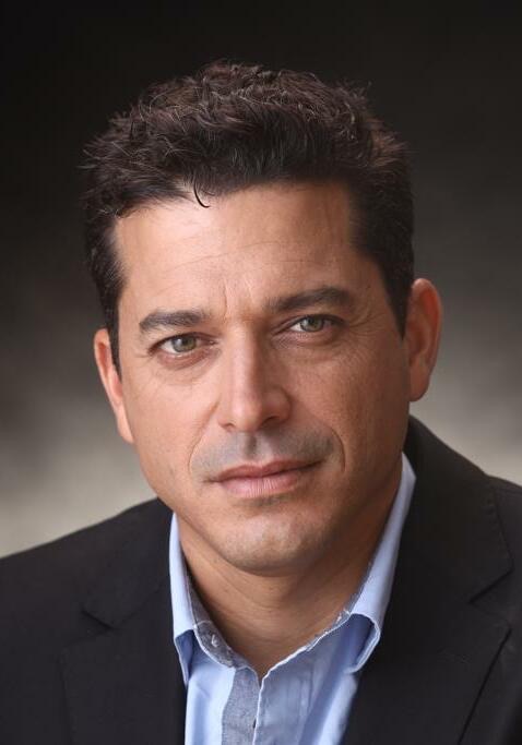 پرتره رسمی نزدیک آمیخای چیکلی، مردی با موهای کوتاه تیره و کت تیره روی پیراهن آبی روشن، رو به دوربین در برابر پس‌زمینه خاکستری تیره، ۲۰۲۲.