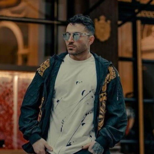 حامد فرد، مرد جوانی با موهای تیره، ته‌ریش کوتاه و عینک آفتابی، با ژاکت تیره روی تی‌شرت سفید، ایستاده در فضای داخلی تزیینی.