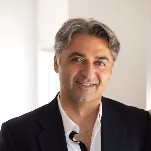 پرتره‌ی هومن خلیلی، مردی با موهایِ تیره‌ی جوگندمی و کتِ مشکی روی پیراهنِ سفیدِ یقه‌باز با میکروفنِ یقه‌ای، که لبخندزنان به یک سو نگاه می‌کند در برابرِ فضایی داخلی و روشن.