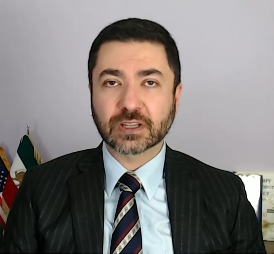 سعید قاسمی‌نژاد، مردی با ریش و کت‌وشلوارِ راه‌راهِ تیره و کراوات، در یک مصاحبه‌ی ویدئویی سخن می‌گوید، در حالی که پرچمِ شیر و خورشیدِ ایران و پرچمِ ایالات متحده پشتِ سرِ اوست، ۲۰۲۵.